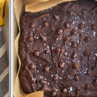Square baking pan with healthy banana brownies sprinkled with sea salt and chocolate chips fresh out of the oven. A fresh banana and a napkin to the sides of the pan.