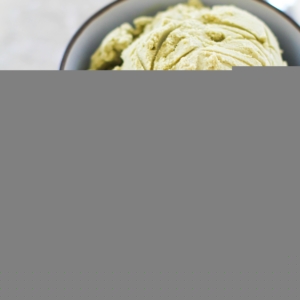 Scoop of green tea ice cream in a bowl with a spoon to the side and a sprinkle of matcha powder to the side.