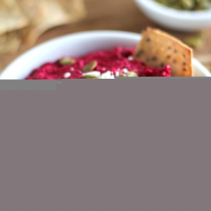 White bowl full of beet hummus with a vibrant red color. Topped with crumbled feta cheese, pumpkin seeds and crackers around for serving.