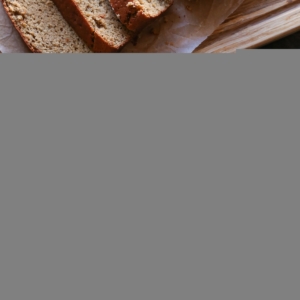 a slice of Almond Butter Banana Bread on a wooden plate with almond butter and strawberry jam spread on top with the rest of the loaf cut into slices on the side and a small bowl of fresh strawberries.
