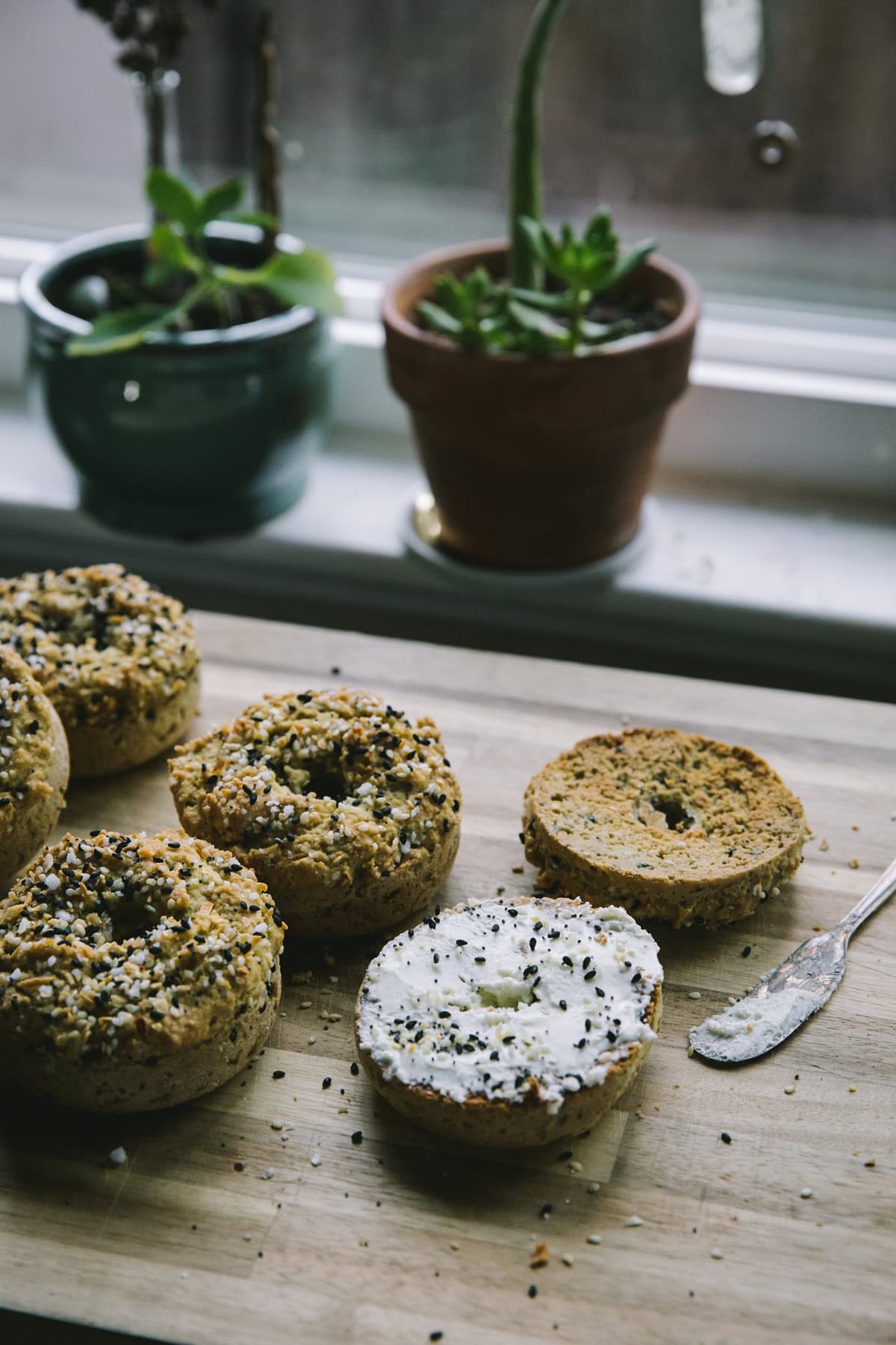 Cutting board with everything bagels on top and one smeared with cream cheese. Plants in the background and a butter knife with cream cheese.