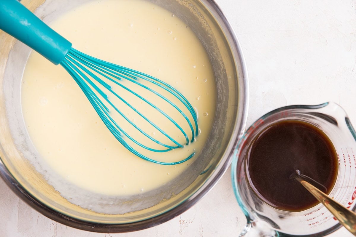Stainless steel bowl of milk, butter, and eggs mixed together with a measuring cup of coffee to the side.