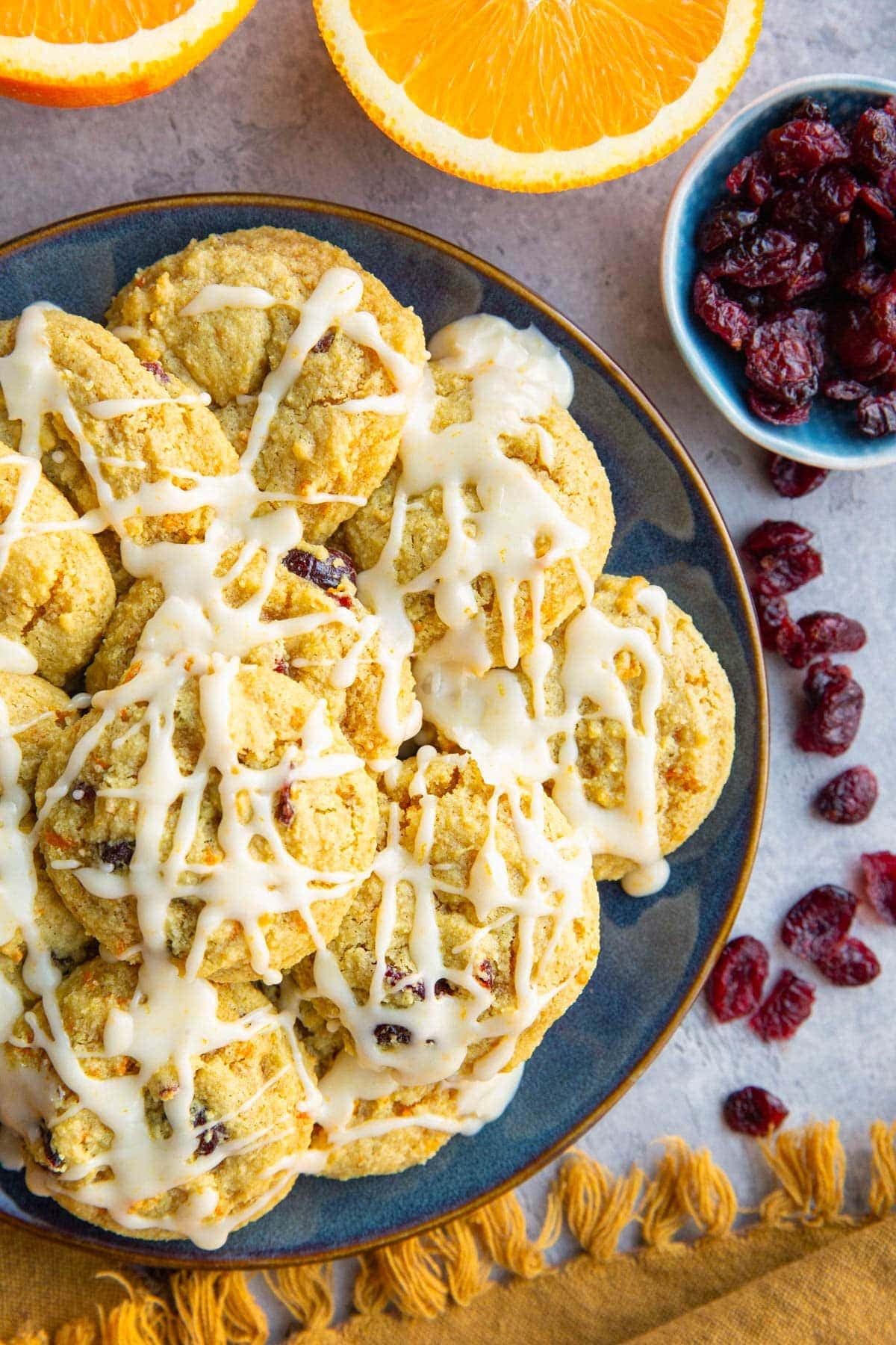 Cranberry orange cookies on a blue plate drizzled with an orange glaze with dried cranberries to the side and a halved orange.