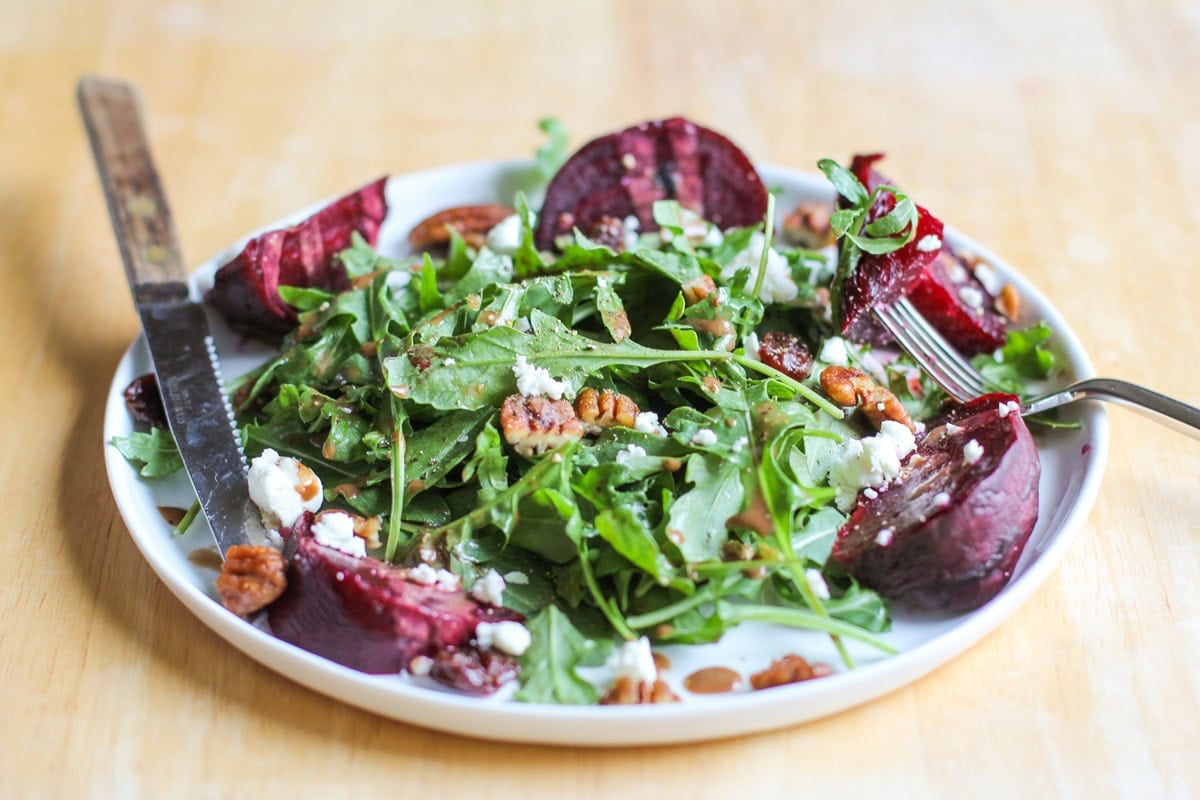 Roasted Beet Arugula Salad with balsamic vinaigrette, pecans, and goat cheese on a white plate with a fork and a knife, ready to eat. A jar of balsamic dressing in the background.