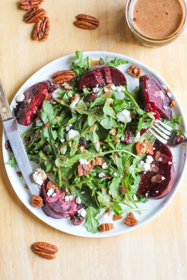 Large white plate with Roasted Beet Salad with Goat Cheese, Arugula, Pecans, dried cranberries and balsamic vinaigrette. Pecans sprinkled around the plate and a jar of dressing to the side.