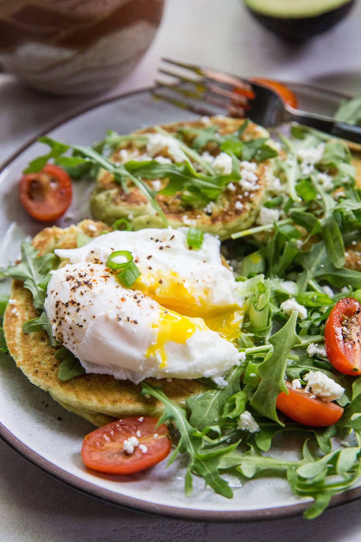 Avocado pancakes on a plate with a poached egg on top and an arugula salad to the side.