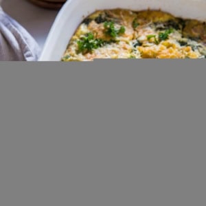 A white baking dish holds a spinach, cheese, and bacon and egg casserole, cut into square pieces and garnished with fresh parsley. Plates, forks, and a striped towel are nearby on a light-colored surface.