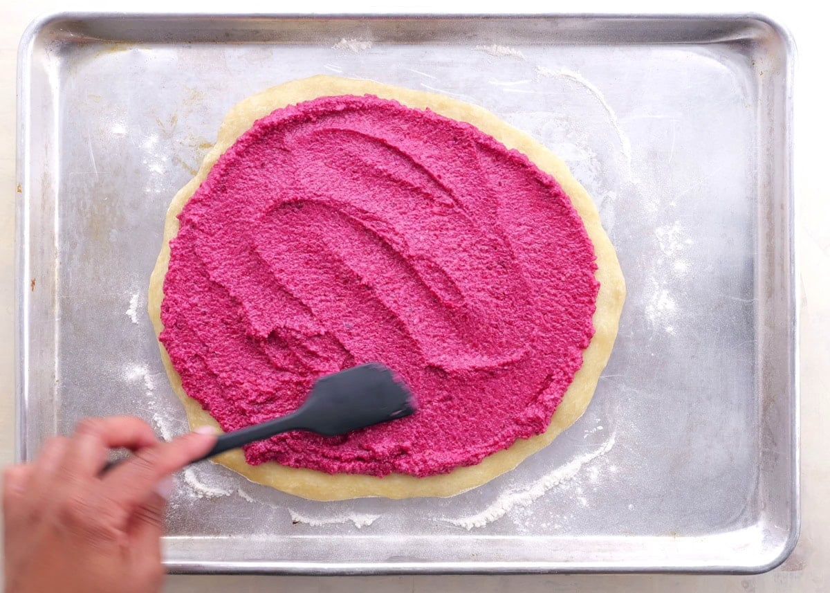Hand spreading beet pesto over pre-baked pizza crust.