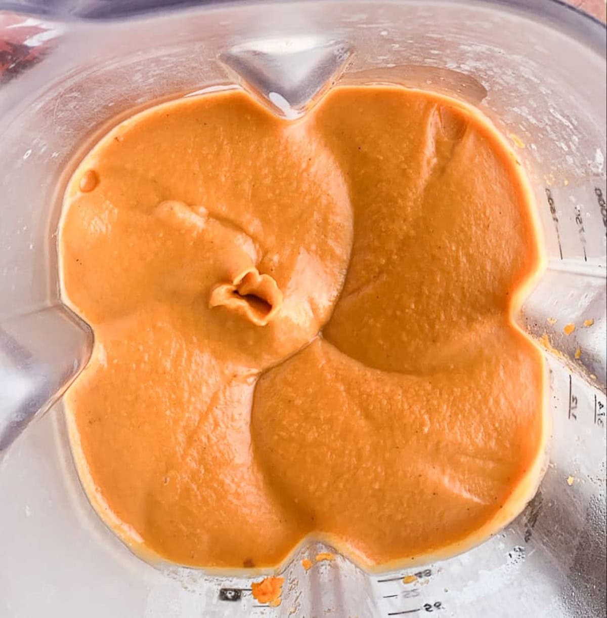 Thick, creamy orange sauce is smoothly blended in a blender, reminiscent of a healthy sweet potato casserole, with visible swirls and ridges on the surface and some residue clinging to the sides.