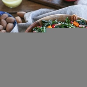Butternut squash kale salad in a large wooden bowl with pecans and pomegranate seeds. A napkin and fresh ingredients all around.