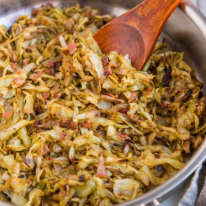 Stainless steel skillet full of caramelized cabbage with onions and bacon. A wooden spoon is ready to serve.