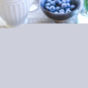 Cauliflower Porridge in a white bowl with milk, honey, fresh berries, and hemp hearts. a mug of tea and a bowl of blueberries in the background.