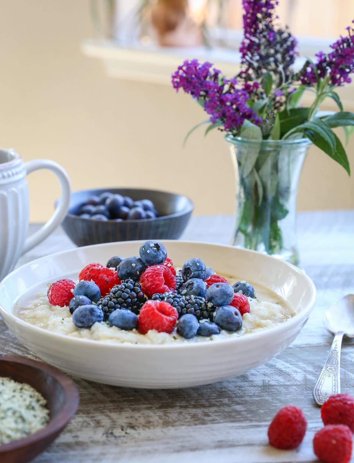 Bowl of Cauliflower Porridge with fresh berries and hemp hearts on top. Fresh berries and a spoon to the side with purple flowers in the background.
