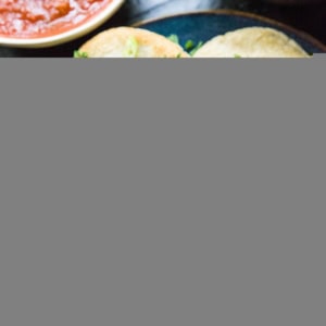 Hand pulling a cheese pupusa apart so you can see the gooey cheesy inside. A plate with more pupusas and a bowl of salsa and beans.