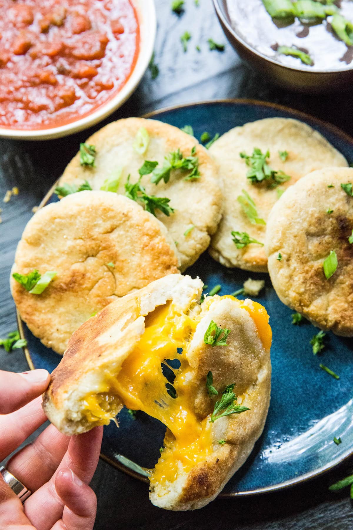 Hand pulling a cheese pupusa apart so you can see the gooey cheesy inside. A plate with more pupusas and a bowl of salsa and beans.