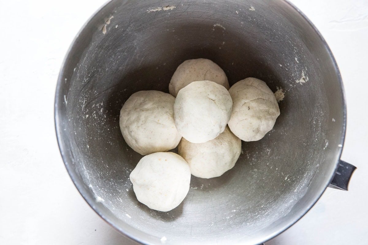 Six balls of dough, ready for shaping into cheese pupusas or a gluten-free cheese flatbread, are placed together inside a large, metal mixing bowl on a white surface.