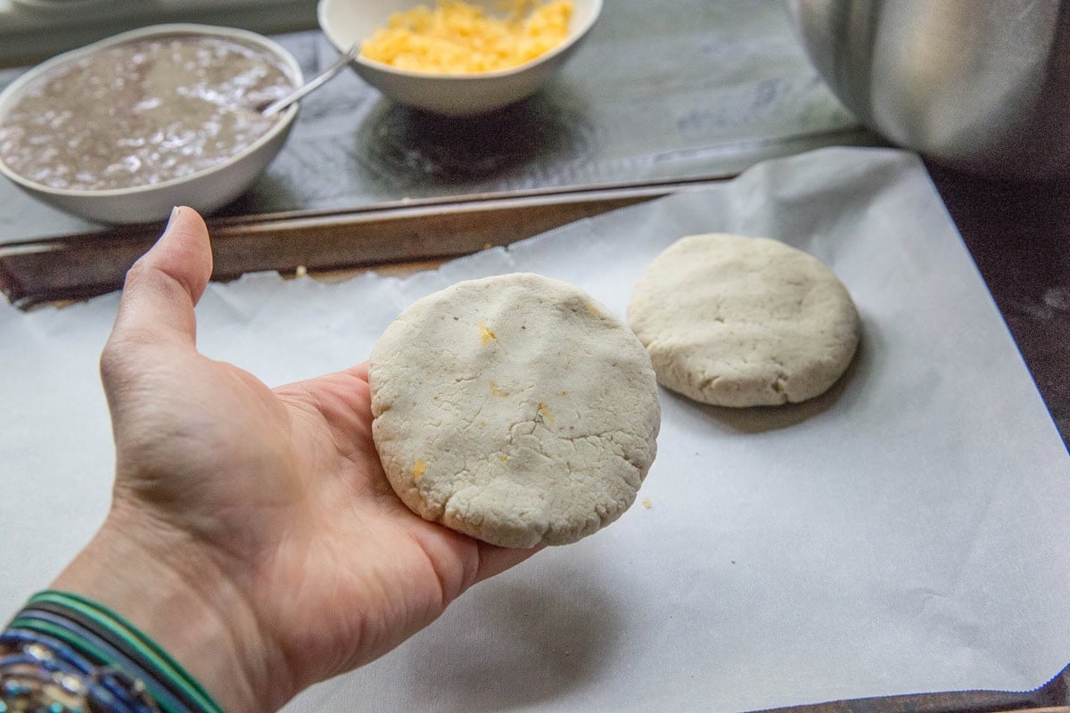 A hand holds a round, uncooked cheese pupusa over a baking sheet lined with parchment paper. Another dough patty and bowls with ingredients for the pupusa recipe are visible in the background.