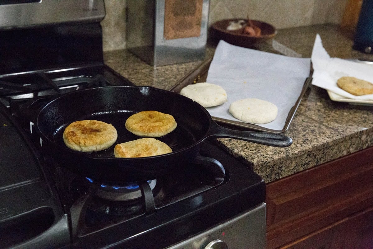 Three round cheese pupusas are sizzling in a cast iron skillet on a gas stove, while two uncooked patties rest on a parchment-lined tray nearby, ready to continue this delicious pupusa recipe.