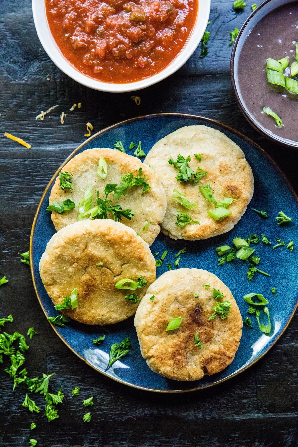 A blue plate with four cheese pupusas, made from a classic pupusa recipe, garnished with chopped green onions and cilantro, sits next to bowls of red salsa and black beans on a dark wooden table.