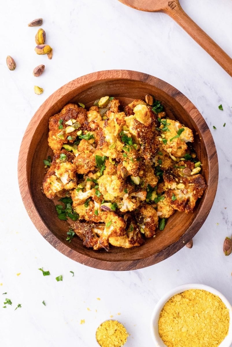 Wooden bowl full of vegan roasted cauliflower with nutritional yeast to the side and fresh parsley sprinkled around.