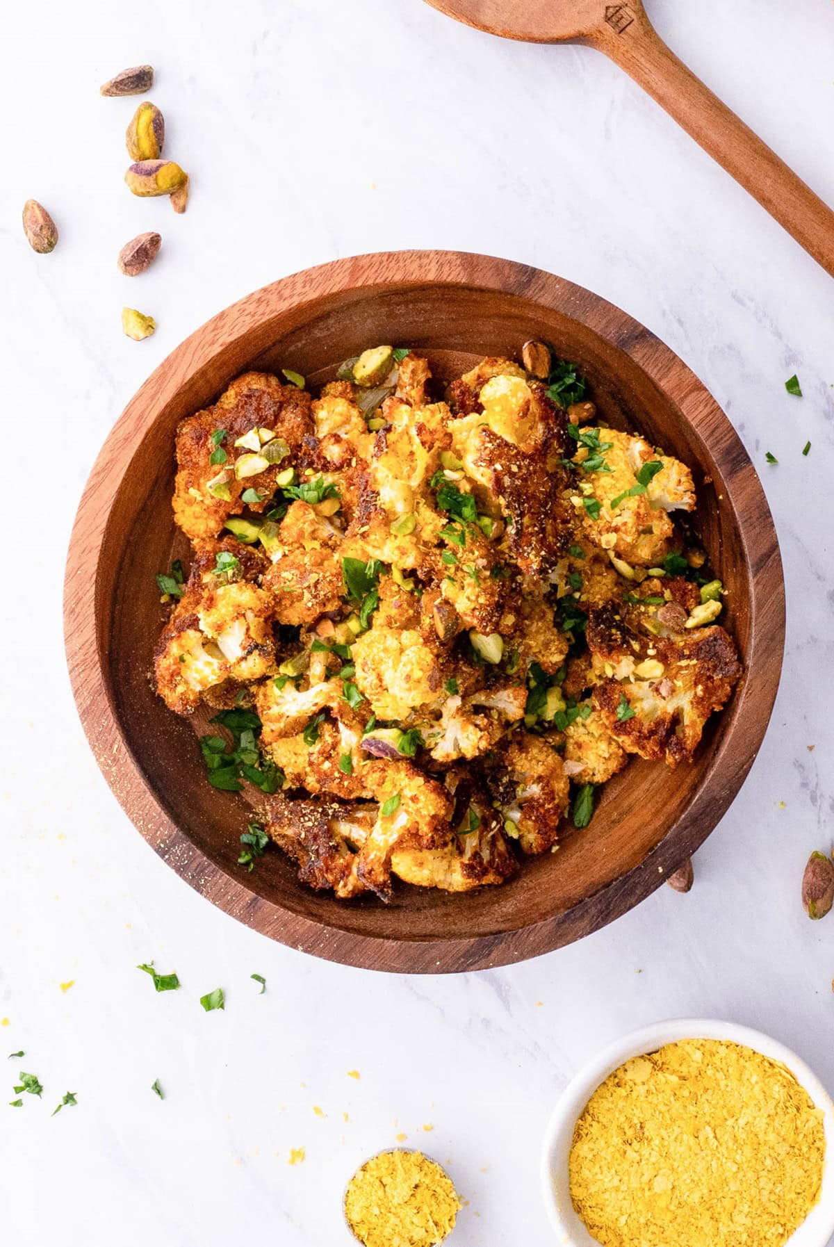 Wooden bowl full of vegan roasted cauliflower with nutritional yeast to the side and fresh parsley sprinkled around.