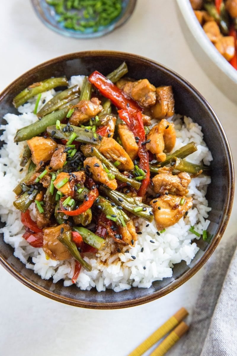 A bowl of white rice topped with Teriyaki Chicken, stir-fried green beans, and red bell peppers, garnished with chopped green onions and black sesame seeds. Chopsticks rest nearby on a light table.