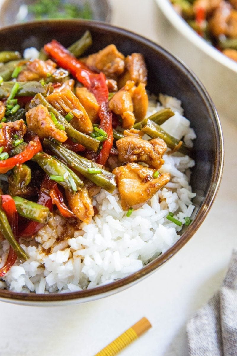 Large blue bowl full of chicken teriyaki on top of white rice, sprinkled with green onions and sesame seeds. Chopsticks and a napkin to the side.
