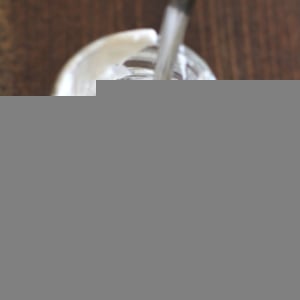 A glass jar filled with creamy white yogurt sits on a wooden surface, topped with a swirl of coconut whipped cream, with a metal spoon inside. A single red cherry lies nearby on the table.