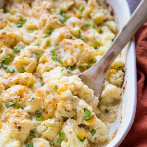 Casserole dish full of cheesy cauliflower casserole with a red napkin to the side and a wooden spoon ready to serve.