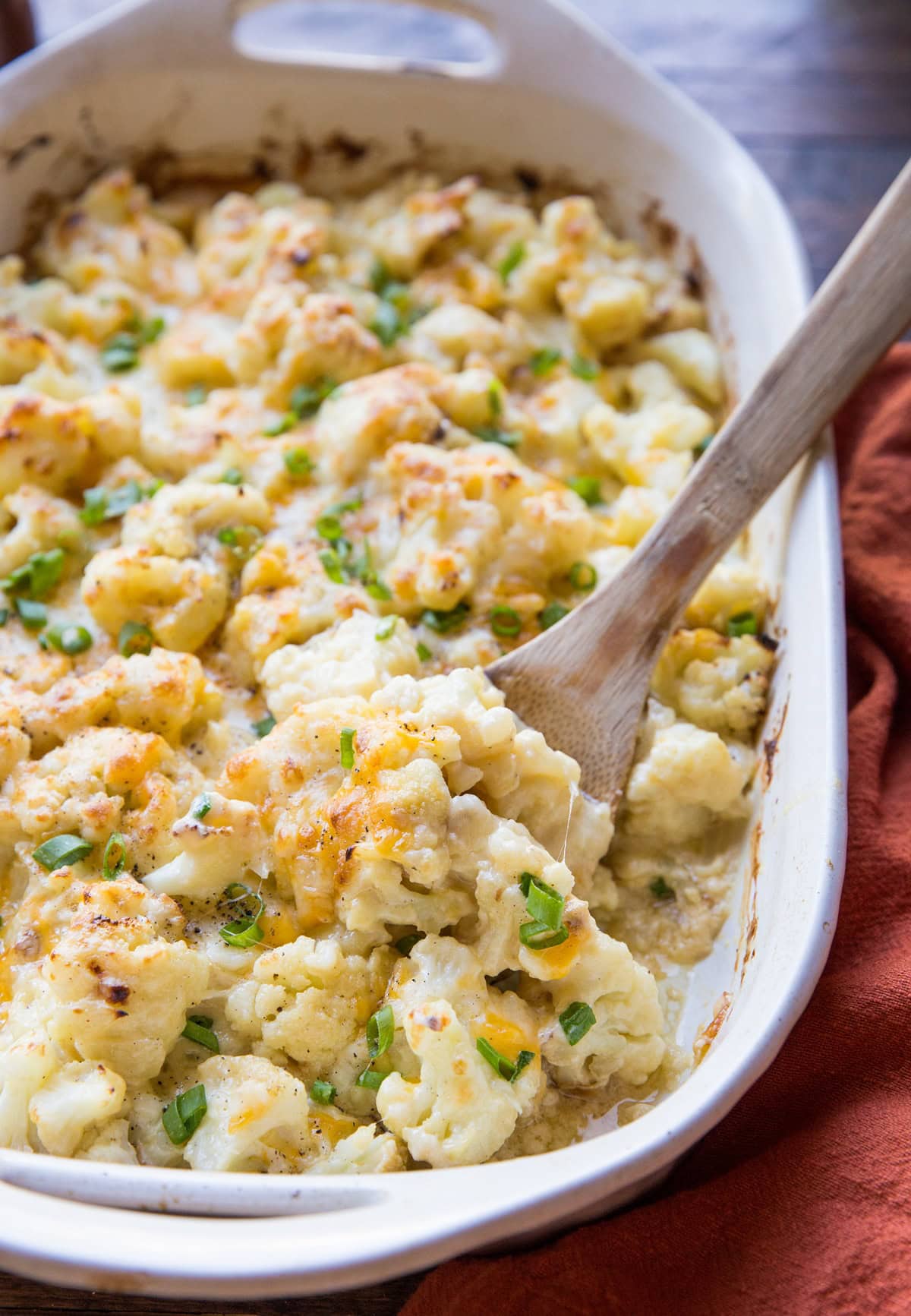 Casserole dish full of cheesy cauliflower casserole with a red napkin to the side and a wooden spoon ready to serve.