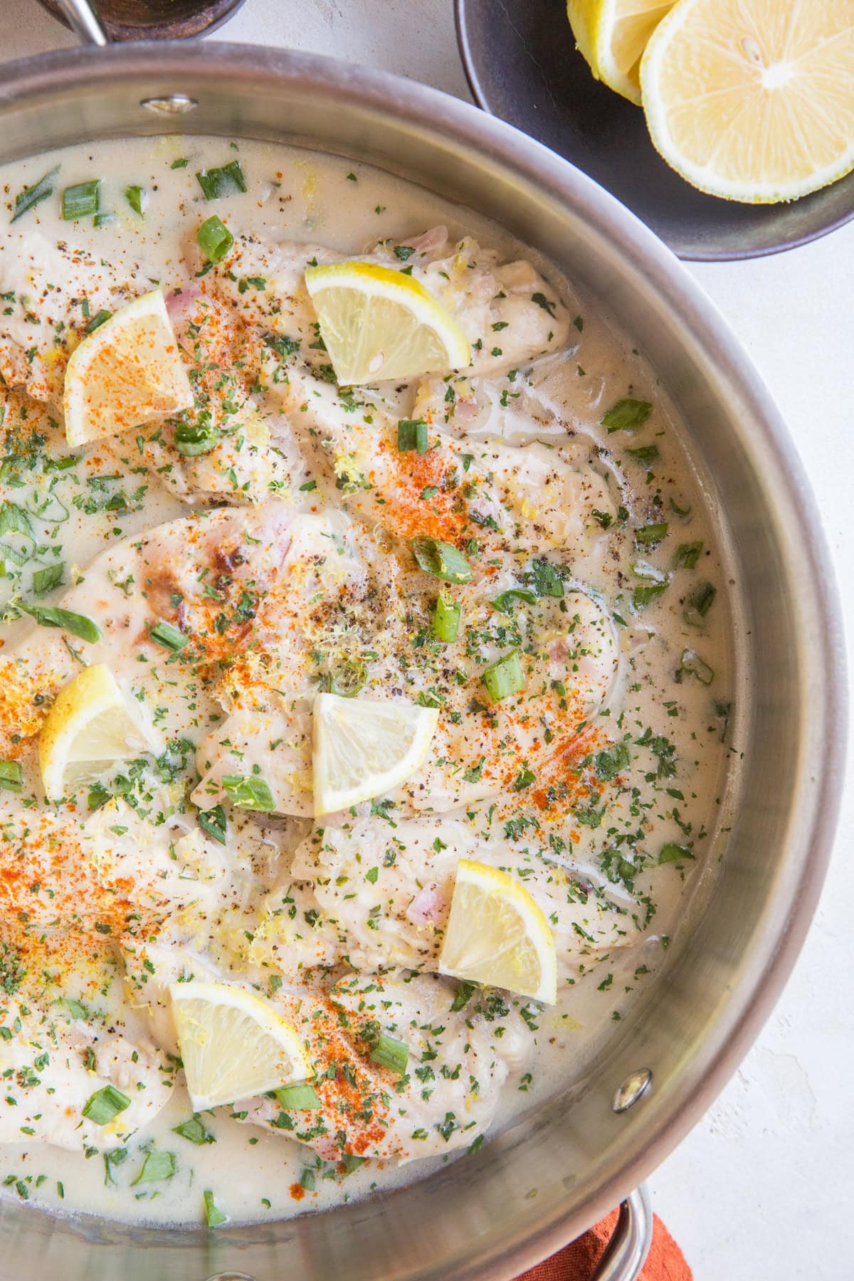 large stainless steel skillet full of chicken in creamy lemon garlic sauce, sprinkled with green onions and dried parley with cut lemon wedges.