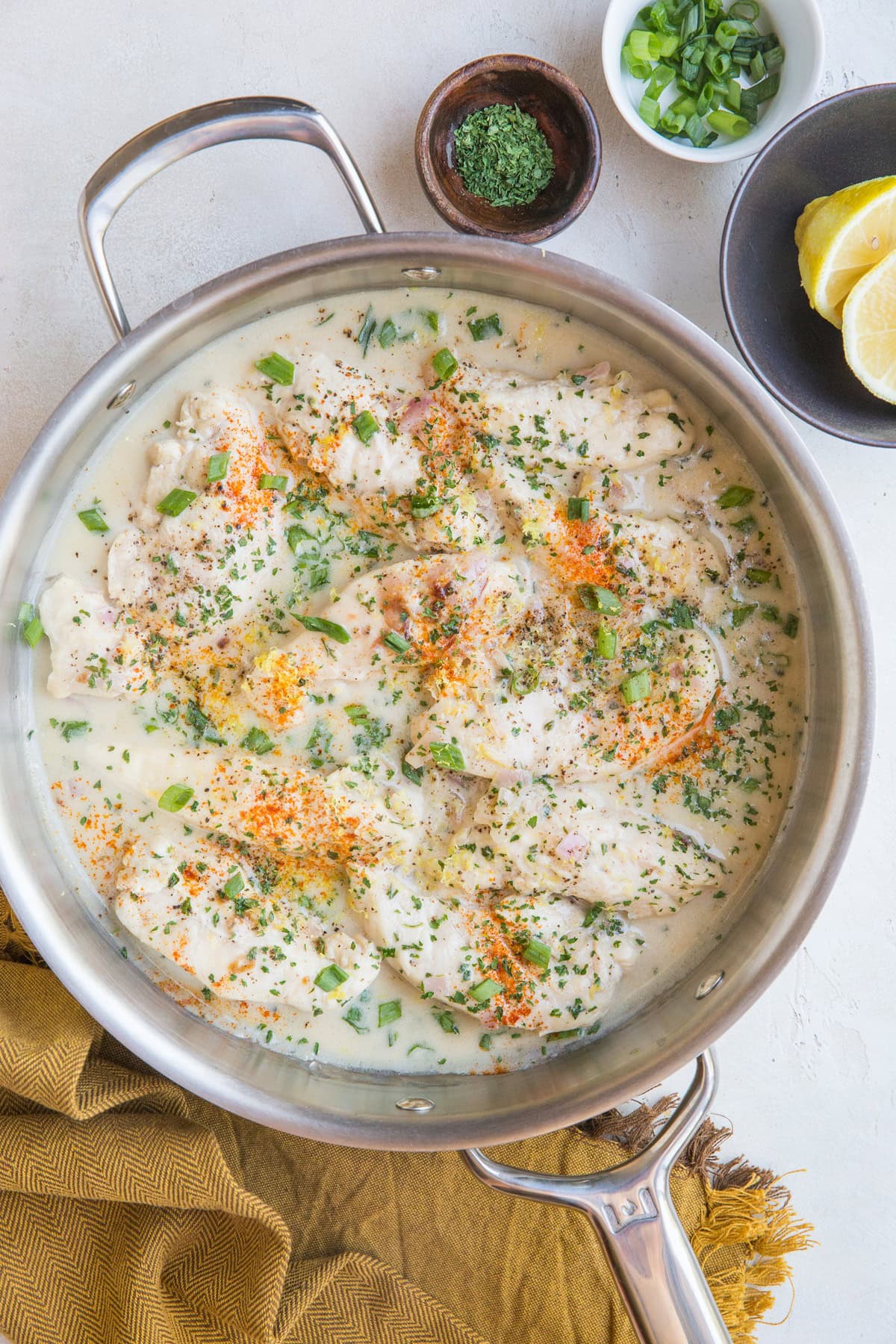 Stainless steel skillet full of chicken in a creamy lemon garlic sauce with a golden napkin and a bowl of fresh lemons to the side.