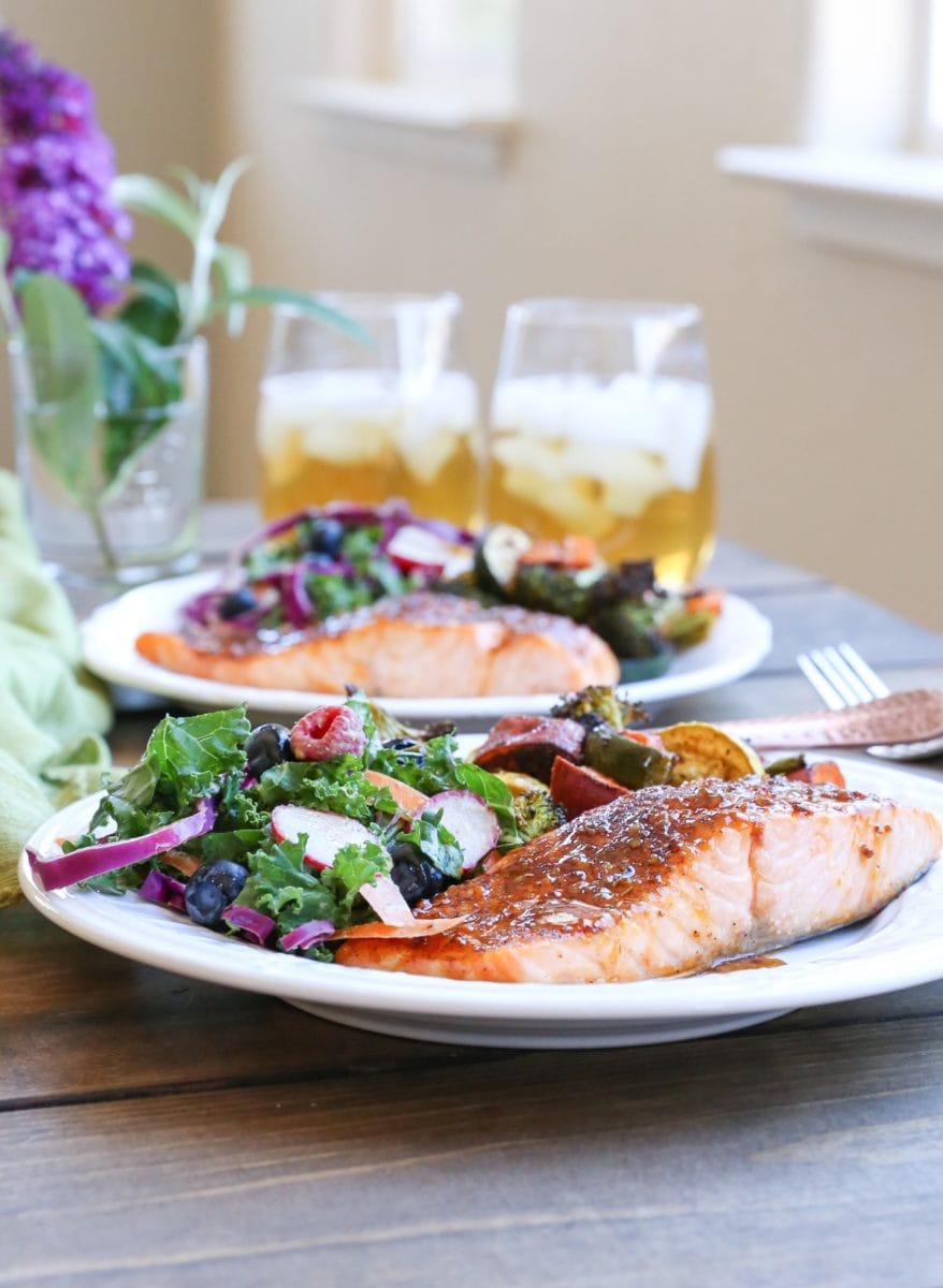 Two plates of Crispy Honey-Glazed Salmon with roasted vegetables and kale salad with glasses of iced tea in the background. Ready to eat.