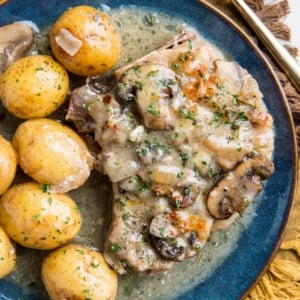 Blue plate with a Pork Chop with creamy garlic sauce, potatoes, and mushrooms with a golden napkin and a gold fork, ready to serve.
