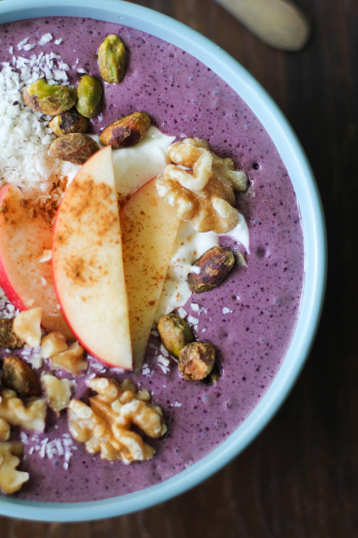 large acai bowl with yogurt, shredded coconut, walnuts, and sliced apples on top inside of a blue bowl.
