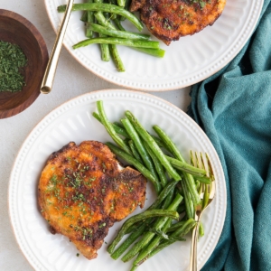 Two plates with cooked air fryer pork chops served with cooked green beans. Golden forks for eating and a blue napkin to the side.