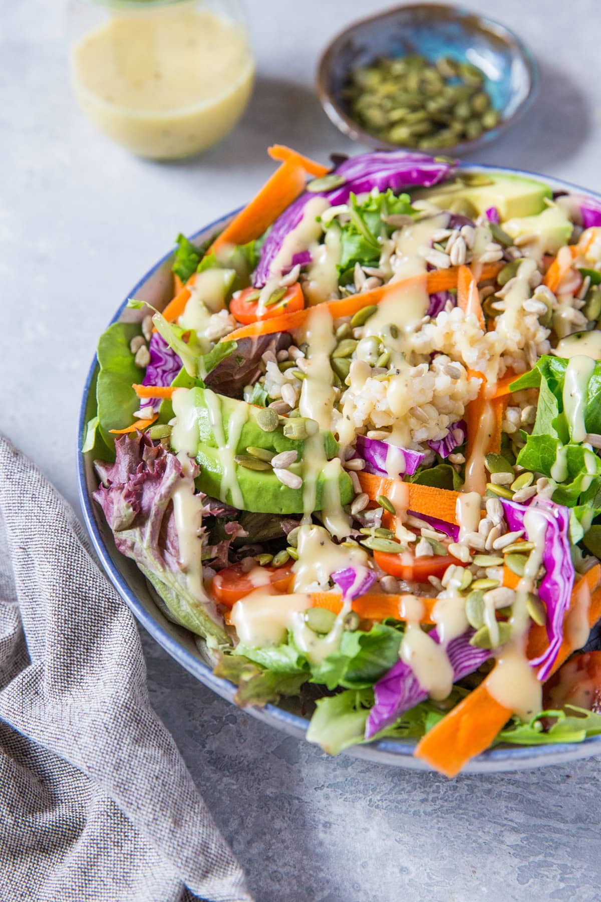 summer salad recipe in a large bowl with greens, carrots, cabbage, avocado, brown rice and homemade dressing. A bowl of pumpkin seeds and a jar of dressing to the side. And a napkin.