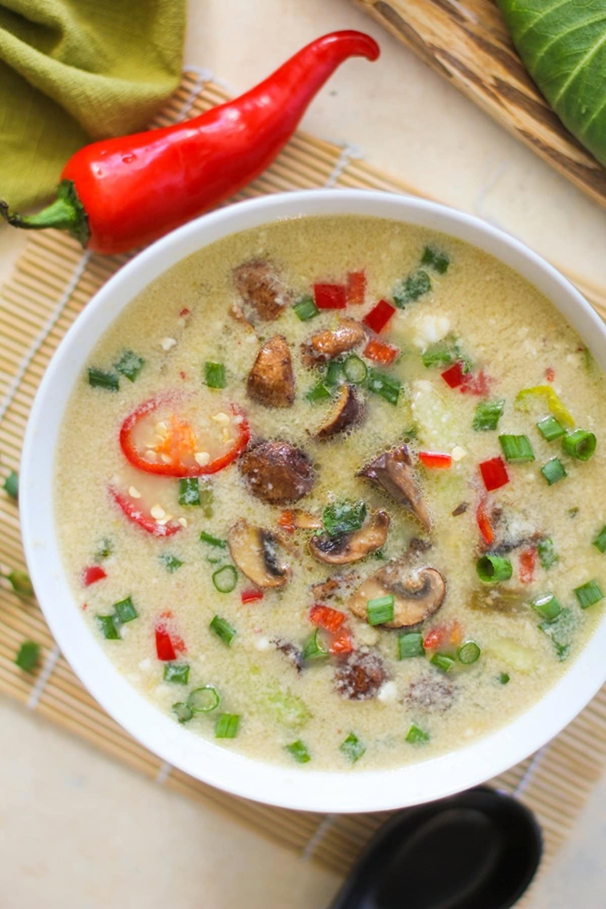 Egg Drop Soup with Bok Choy and Mushrooms sitting on a white backdrop with a soup spoon to the side. The soup is sprinkled with green onions and sauteed mushrooms.