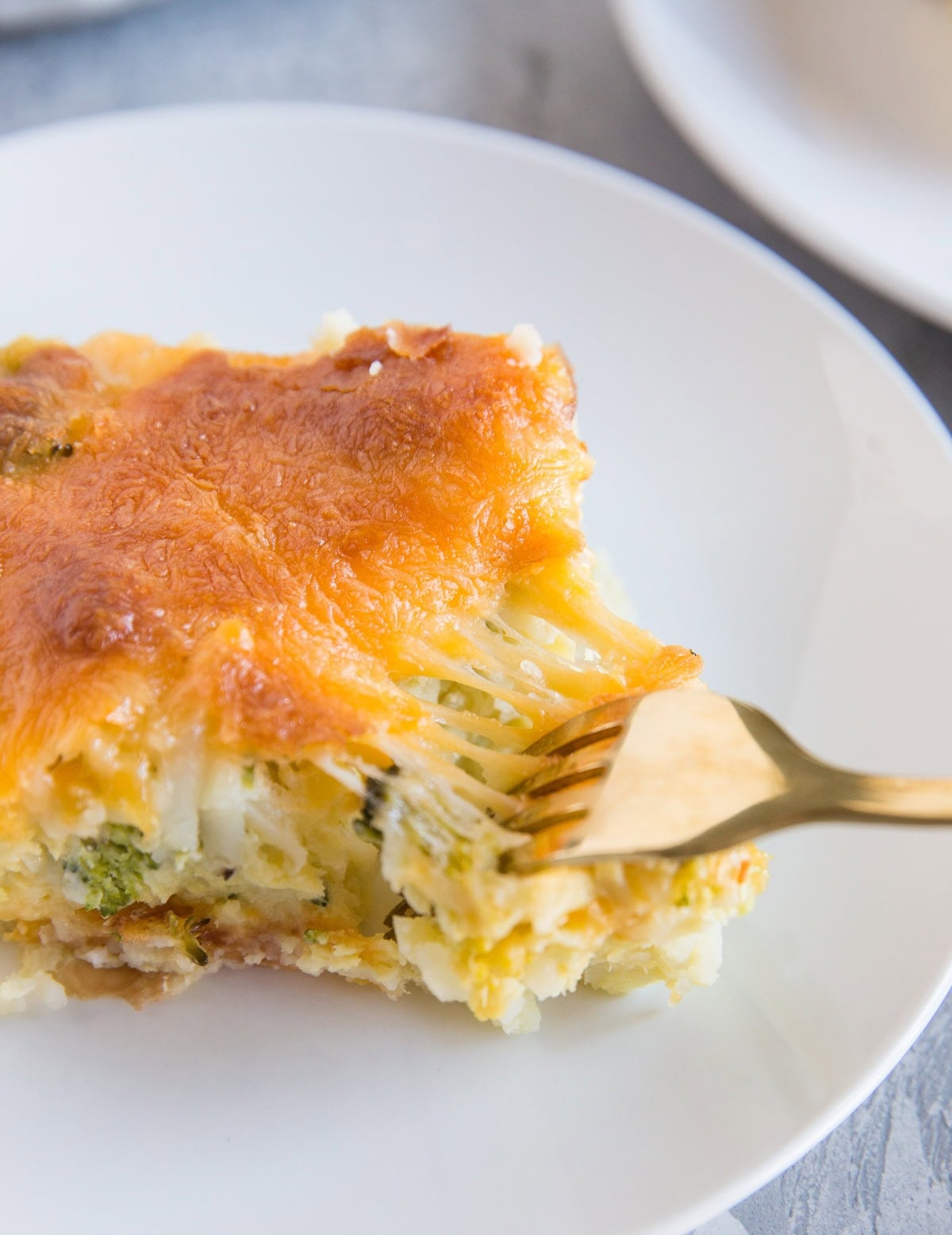 Slice of cheddar bacon egg hashbrown casserole on a plate with a fork pulling at a piece to reveal a cheese pull.