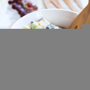 White bowl of fruit salad with a bunch of grapes in the background and a fresh basil leaf in front of the bowl.