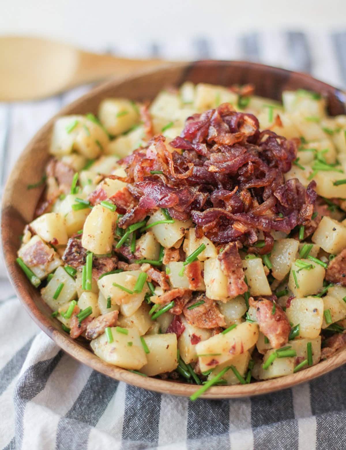 German-Style Potato Salad with bacon, caramelized onions, and chives in a bowl with the caramelized onions on top. The wooden bowl is on top of a striped napkin and the dish is ready to serve.