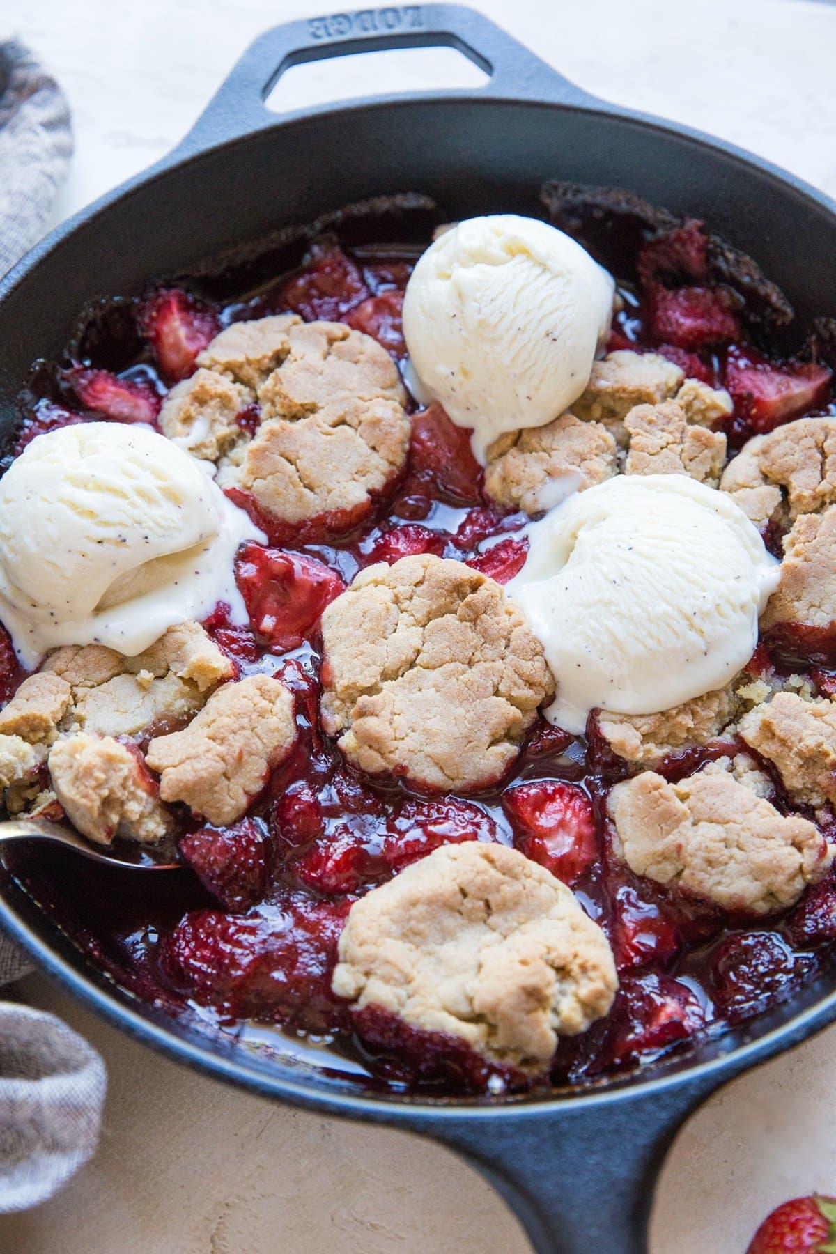 Cast iron skillet full of strawberry cobbler with a scoop of ice cream and a serving spoon, ready to serve.
