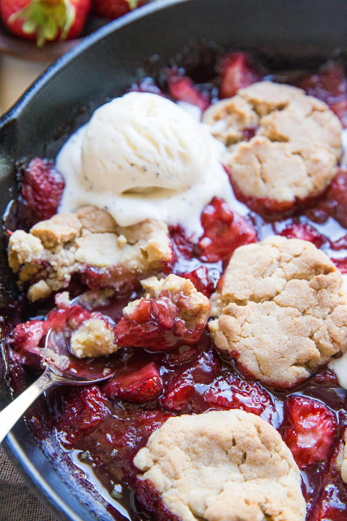 Large skillet full of strawberry cobbler with a scoop of melted vanilla ice cream and a spoon scooping out some of the cobbler.