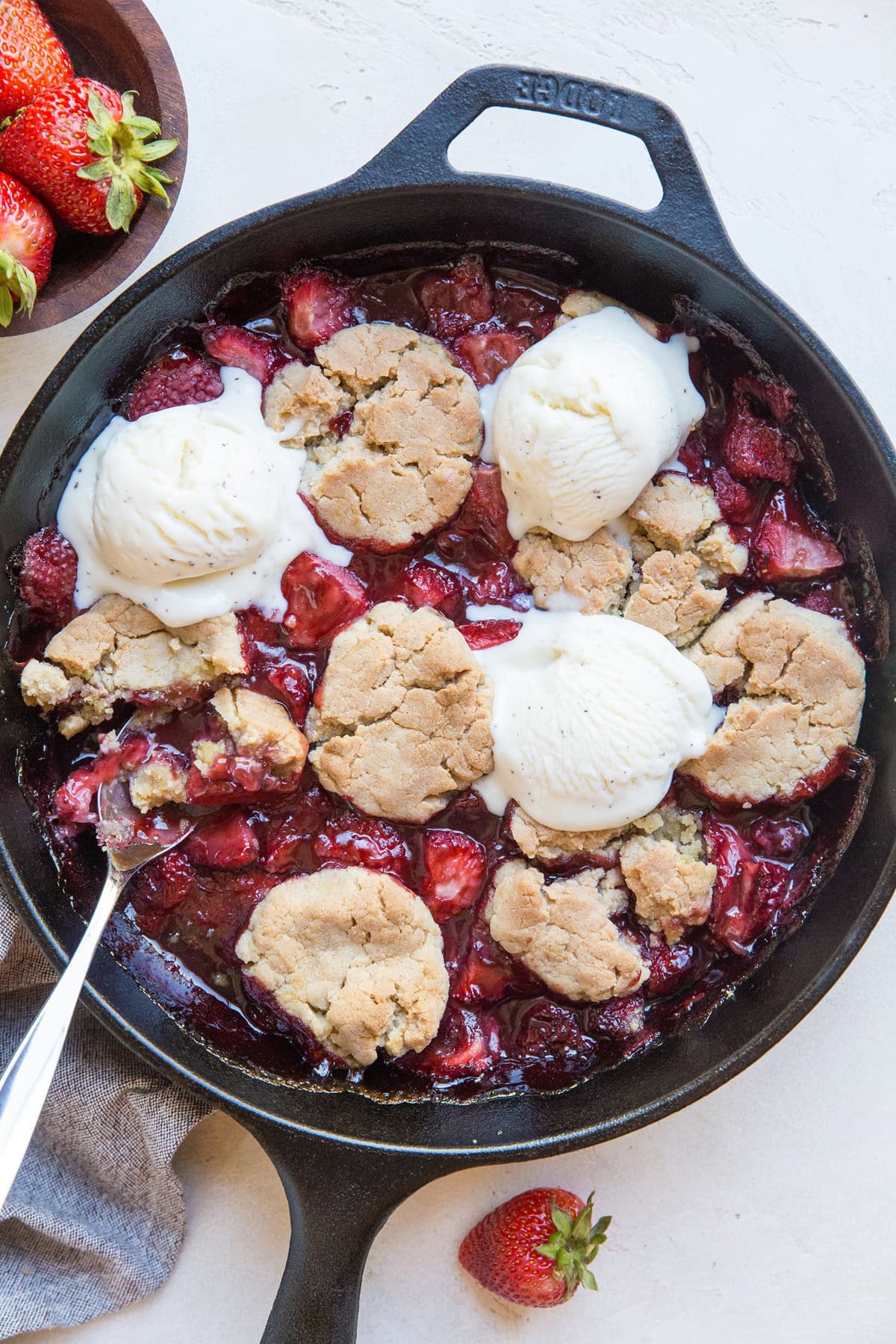 Cast iron skillet full of strawberry cobbler with scoops of vanilla ice cream on top and fresh strawberries to the side.