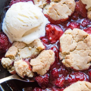 Strawberry cobbler in a large cast iron skillet with scoops of melted ice cream on top and a spoon scooping out some of the cobbler.