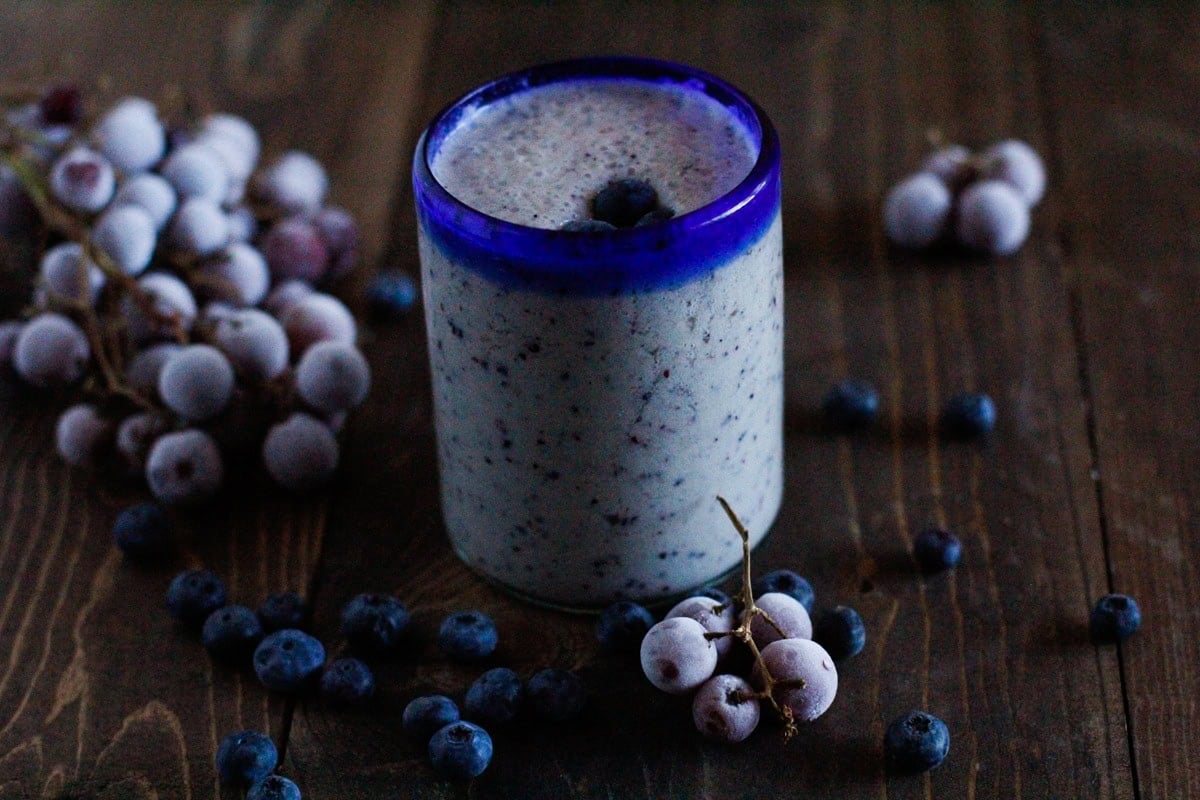 Glass of grape smoothie on a wooden table with frozen grapes and fresh blueberries to the side.
