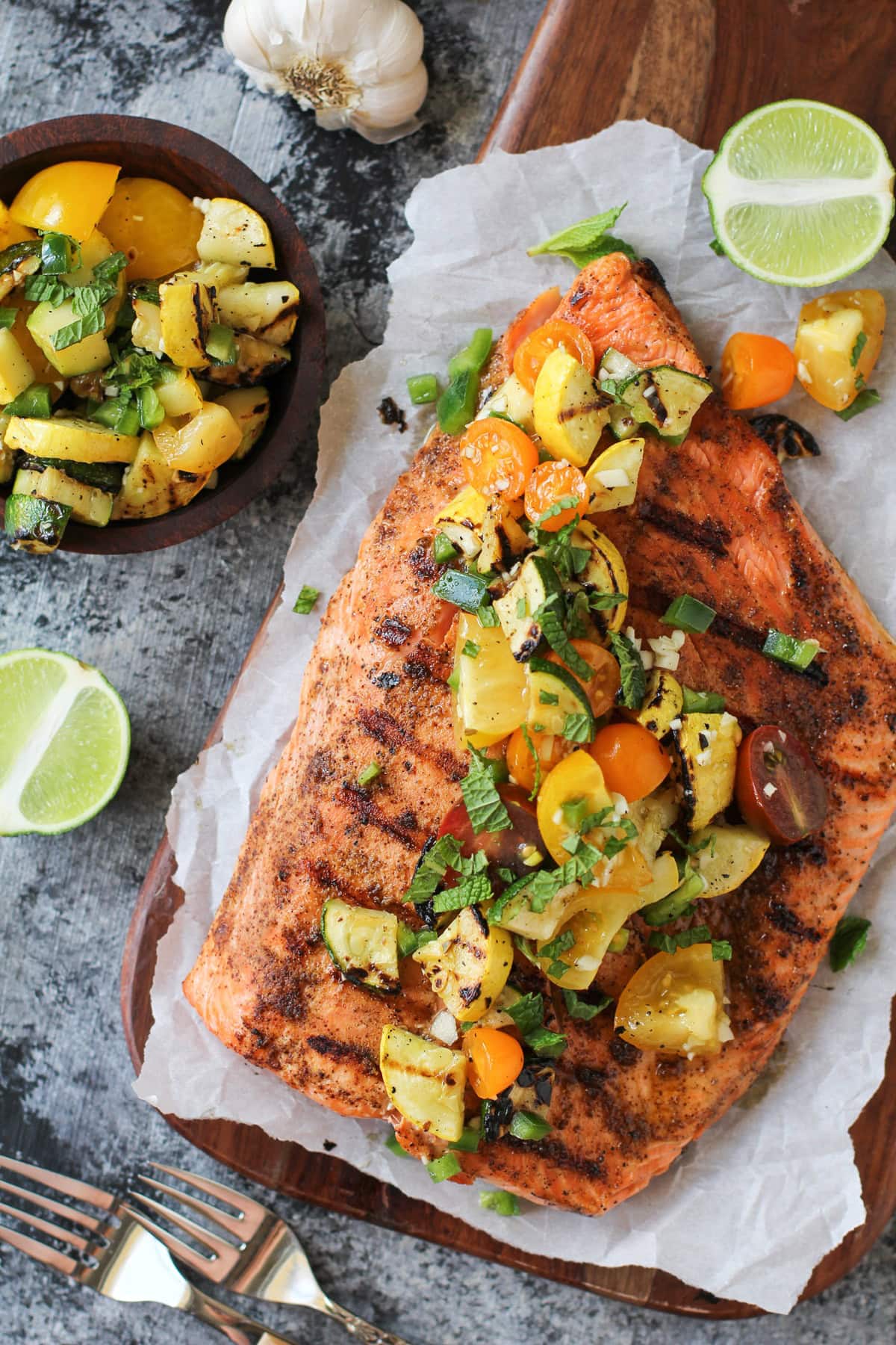 How to Perfectly Grill Salmon - the best grilled salmon on a cutting board with grilled zucchini and tomatoes on top, with sprinkles of green onions and fresh mint leaves.