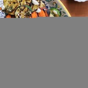 Platter of grilled sweet potatoes, yellow squash, and zucchini with feta cheese sprinkled on top and pesto sauce drizzled over everything.