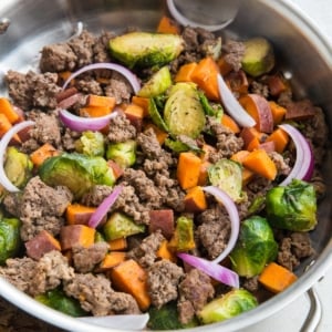 large stainless steel skillet with cooked ground beef, sweet potatoes, and brussel sprouts.