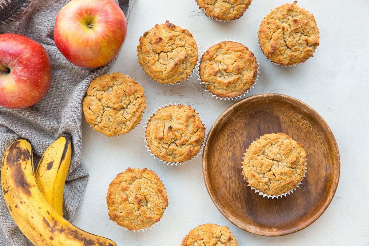 Healthy Apple Cinnamon Muffins on a white table with ripe bananas and fresh apples to the side with a grey napkin.